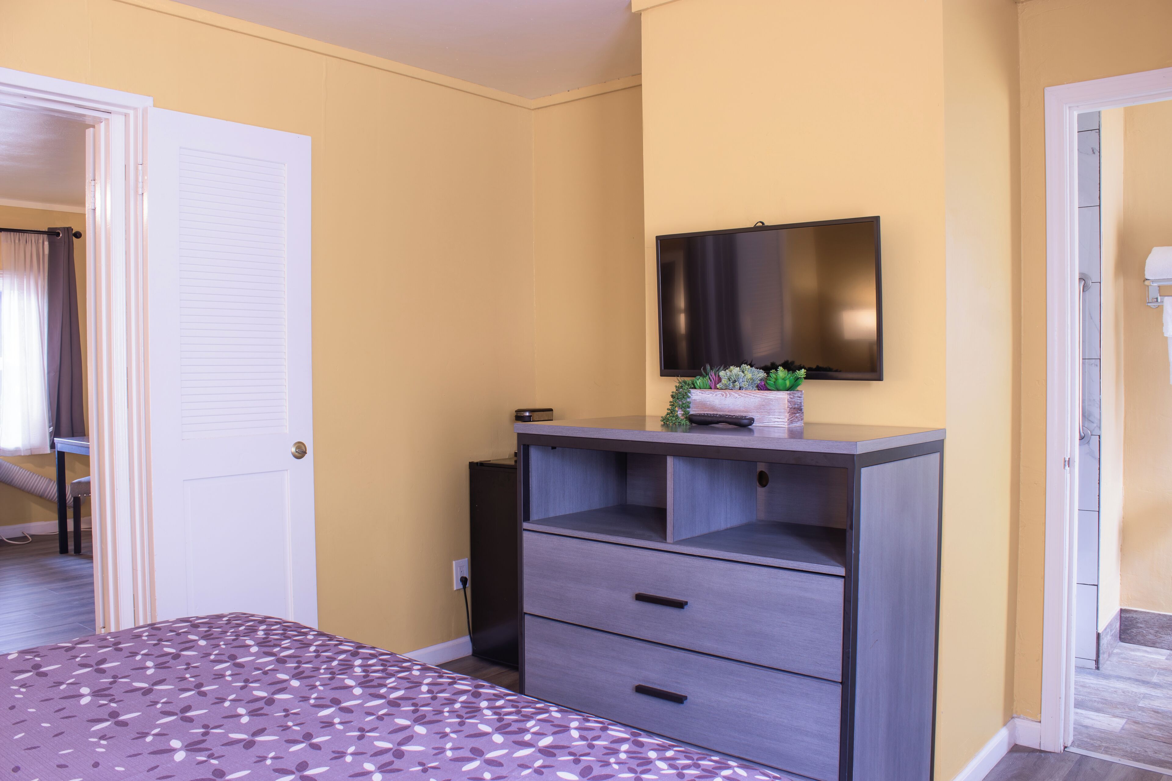 A bedroom with a bed, a dresser, a television mounted on the wall, and a door leading to another room.