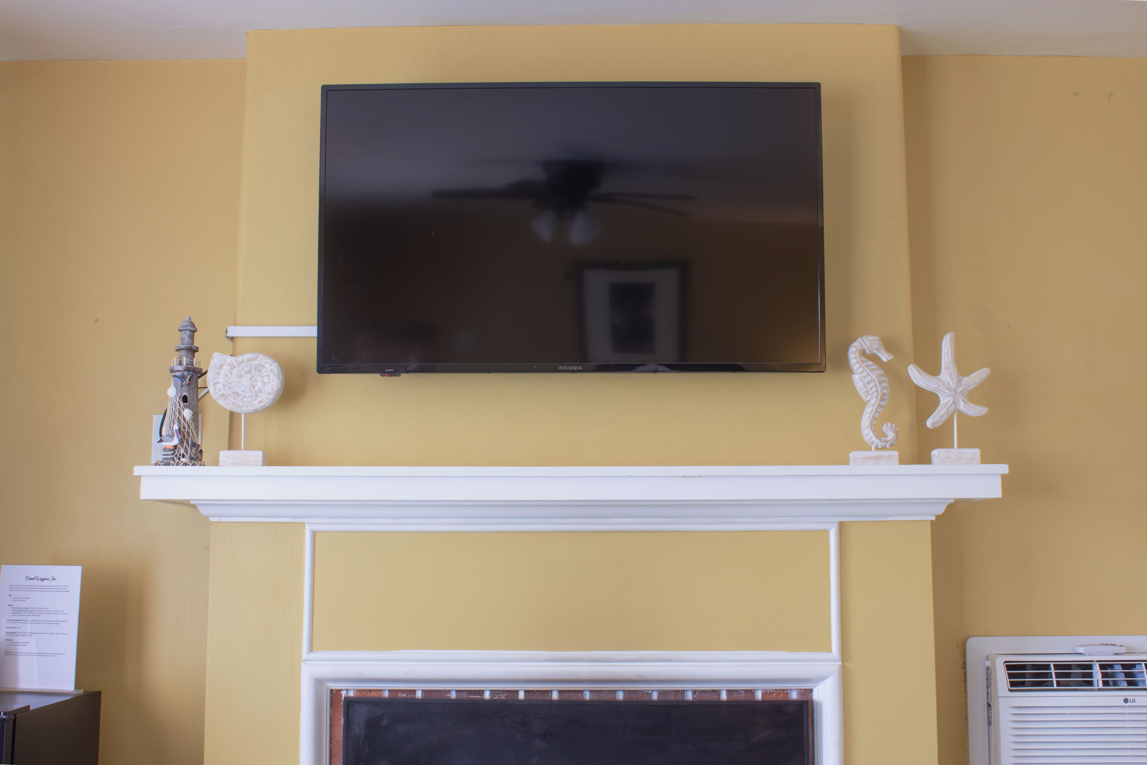 A living room with a mounted flat-screen TV above a white mantelpiece, a fireplace below, and decorative items on the mantel.