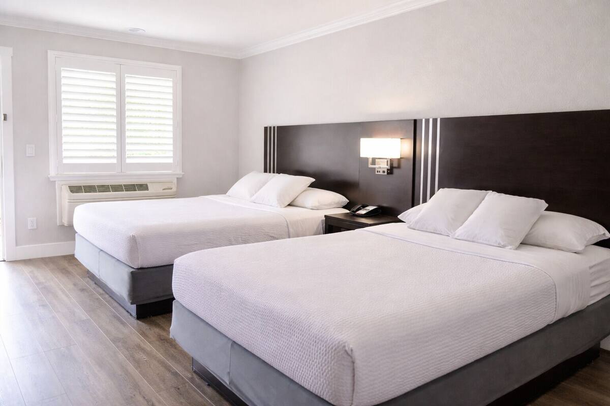 A hotel room with two beds, a nightstand with a phone, a wall-mounted lamp, and a window with blinds.