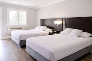A hotel room with two beds, a nightstand with a phone, a wall-mounted lamp, and a window with blinds.