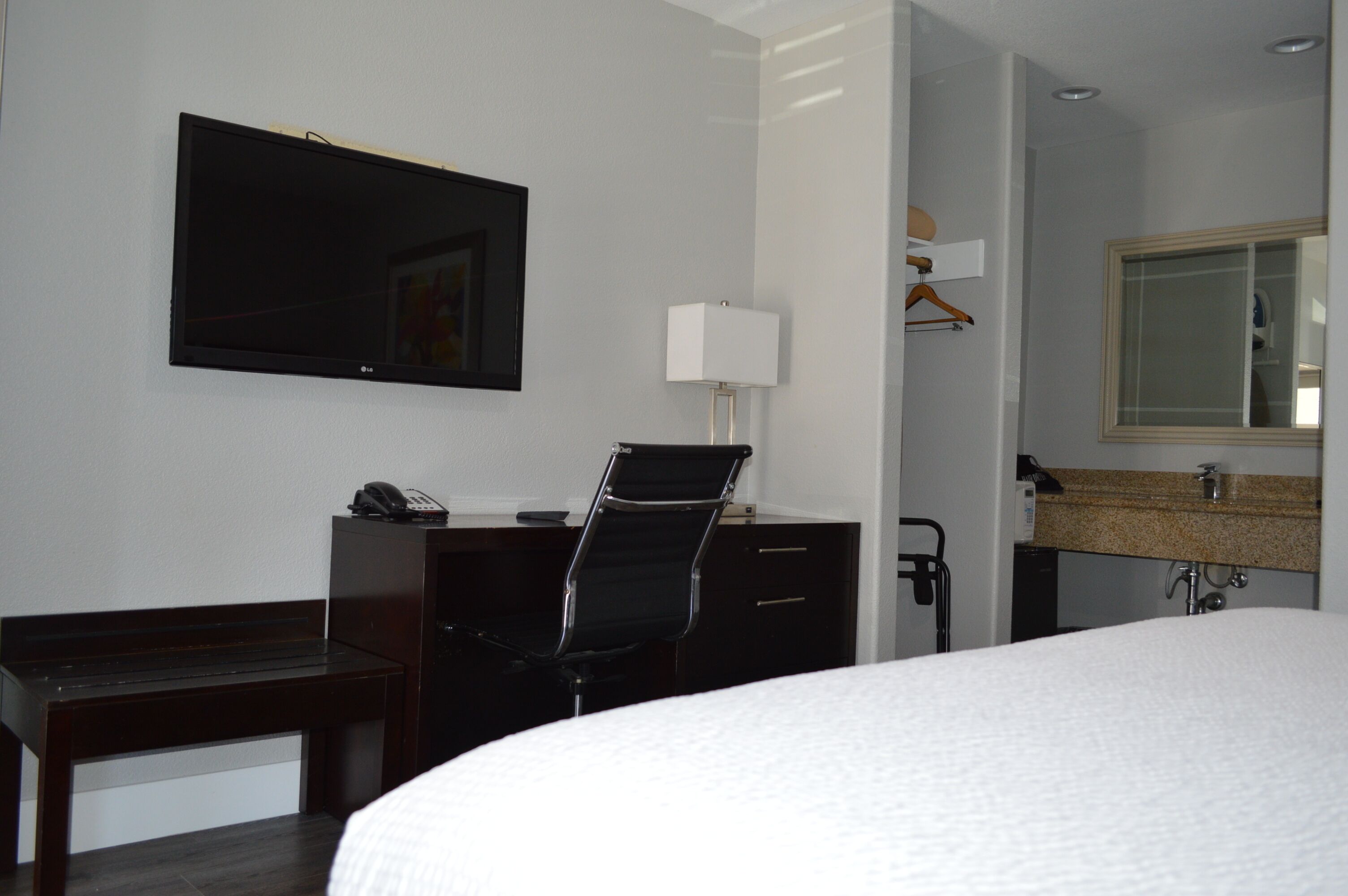A hotel room with a bed, a desk with a chair, a television, a lamp, a mirror, and a bathroom with a sink and a shower.
