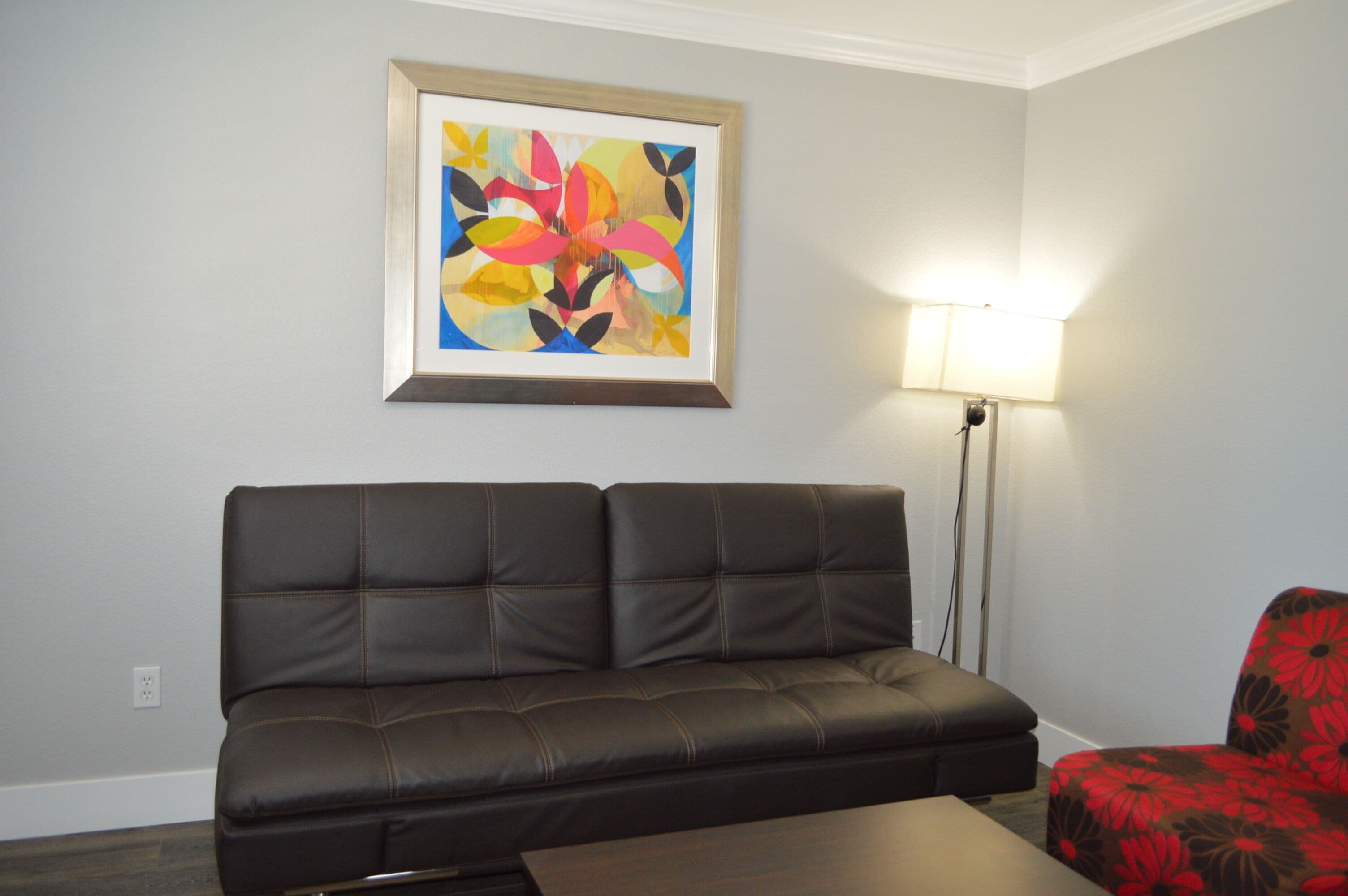 Un salón con un sofá de cuero marrón oscuro, un sillón floral rojo y negro, una lámpara de pie y un cuadro abstracto colorido en la pared.
