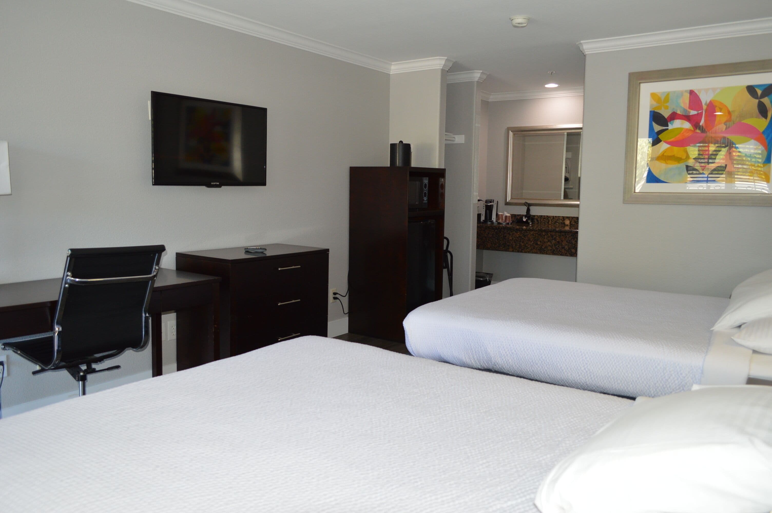 A hotel room with two beds, a desk, a chair, a TV, and a bathroom visible in the background.