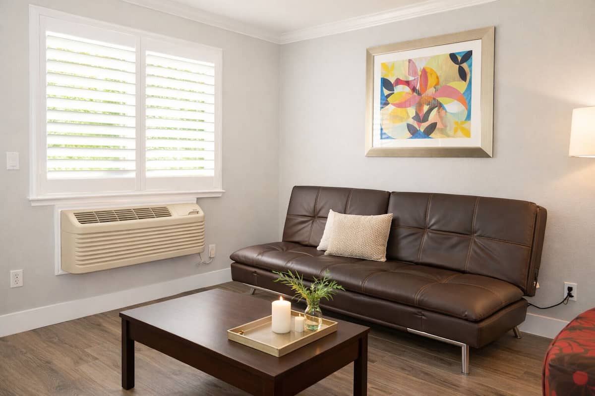 A living room with a brown leather sofa, a wooden coffee table with a candle and a plant, a window with blinds, and a colorful abstract painting on the wall.