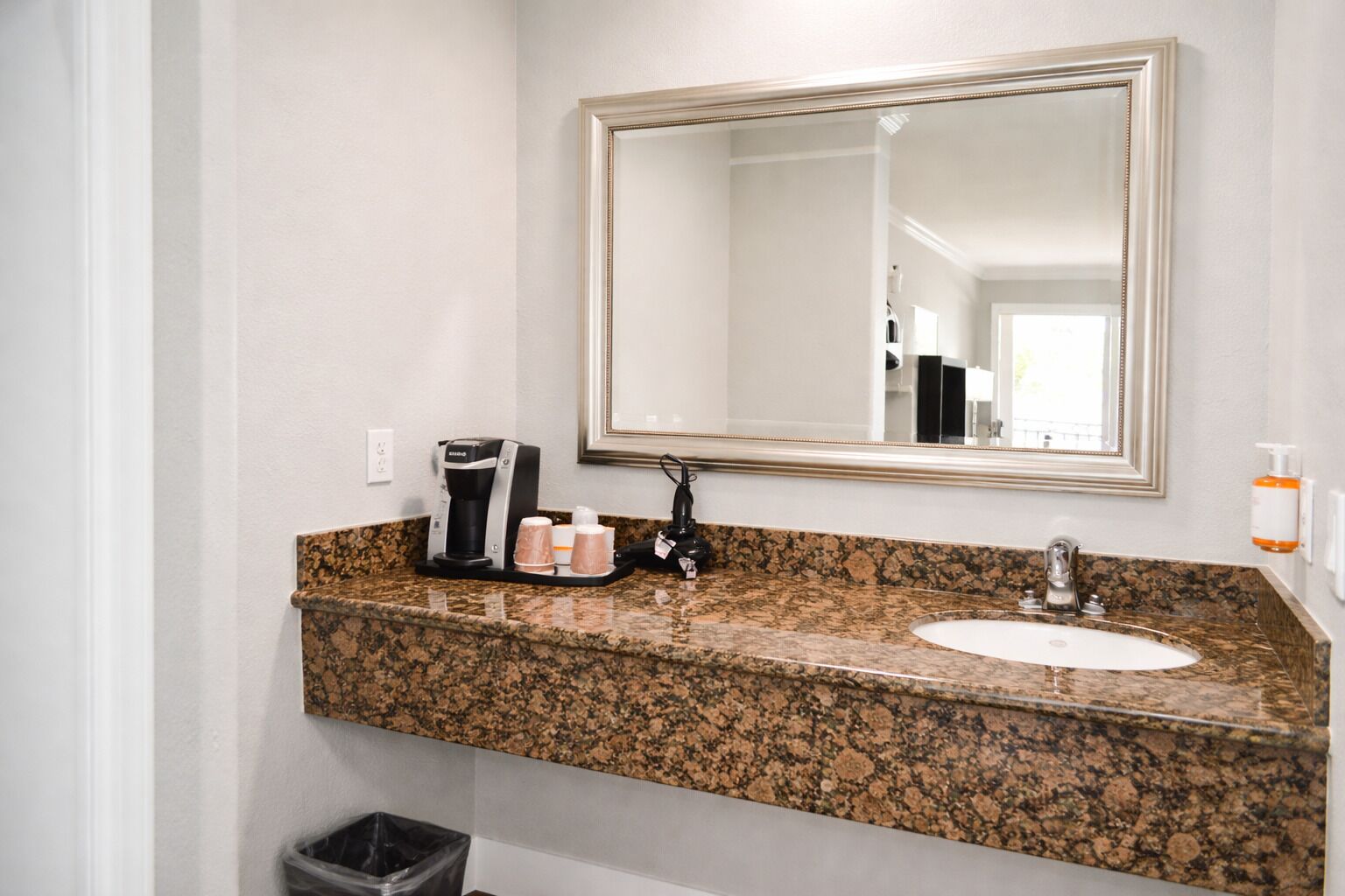 A bathroom with a granite countertop, a white sink, a coffee maker, and a trash can.