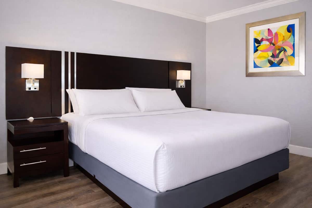 A hotel room with a large bed, two bedside tables, a nightstand, a wall-mounted painting, and a wooden floor.