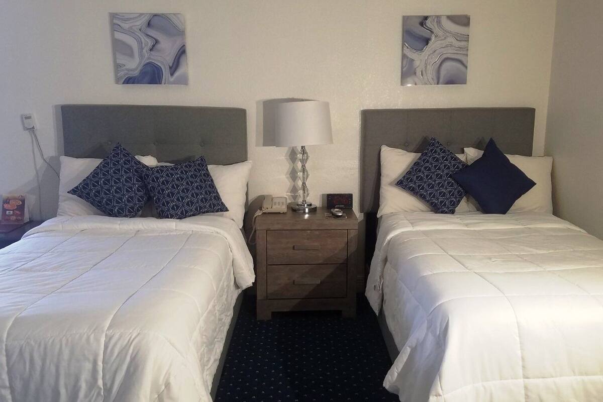 Et hotelværelse med to senge, hver med hvide sengetæpper og puder med geometriske mønstre. Et natbord med lampe, telefon og et lille ur står imellem sengene. Værelset har et enkelt og rent design med mørkt tæppe og abstrakt kunst på væggene.
