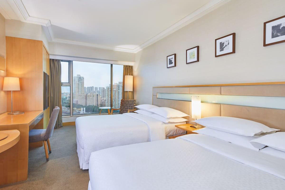 A hotel room with two beds, a desk, a chair, and a TV. There are framed pictures on the wall and a view of the city through large windows.