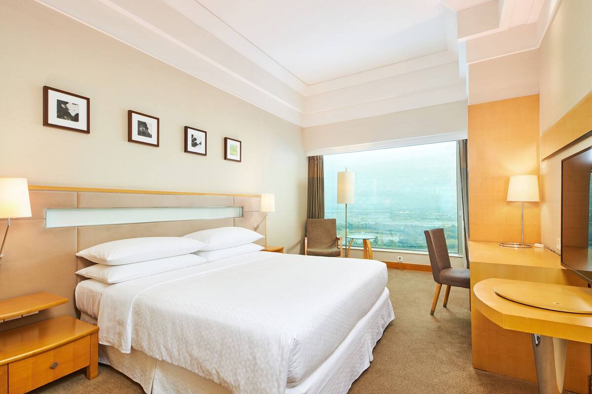 A hotel room with a large bed, bedside tables, a desk, and a chair. There are framed pictures on the wall and a large window with a view.