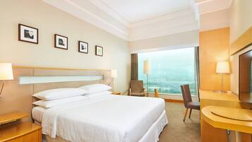 A hotel room with a large bed, bedside tables, a desk, and a chair. There are framed pictures on the wall and a large window with a view.