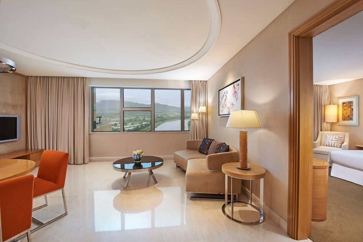A modern hotel room with a large window, a sofa, a round coffee table, and a TV.