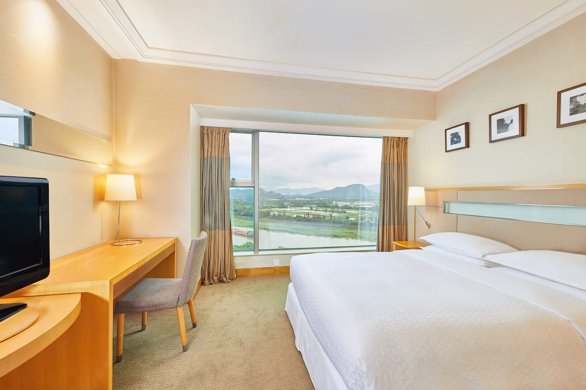A hotel room with a large bed, a desk with a TV, a chair, a nightstand, and a view of mountains through the window.