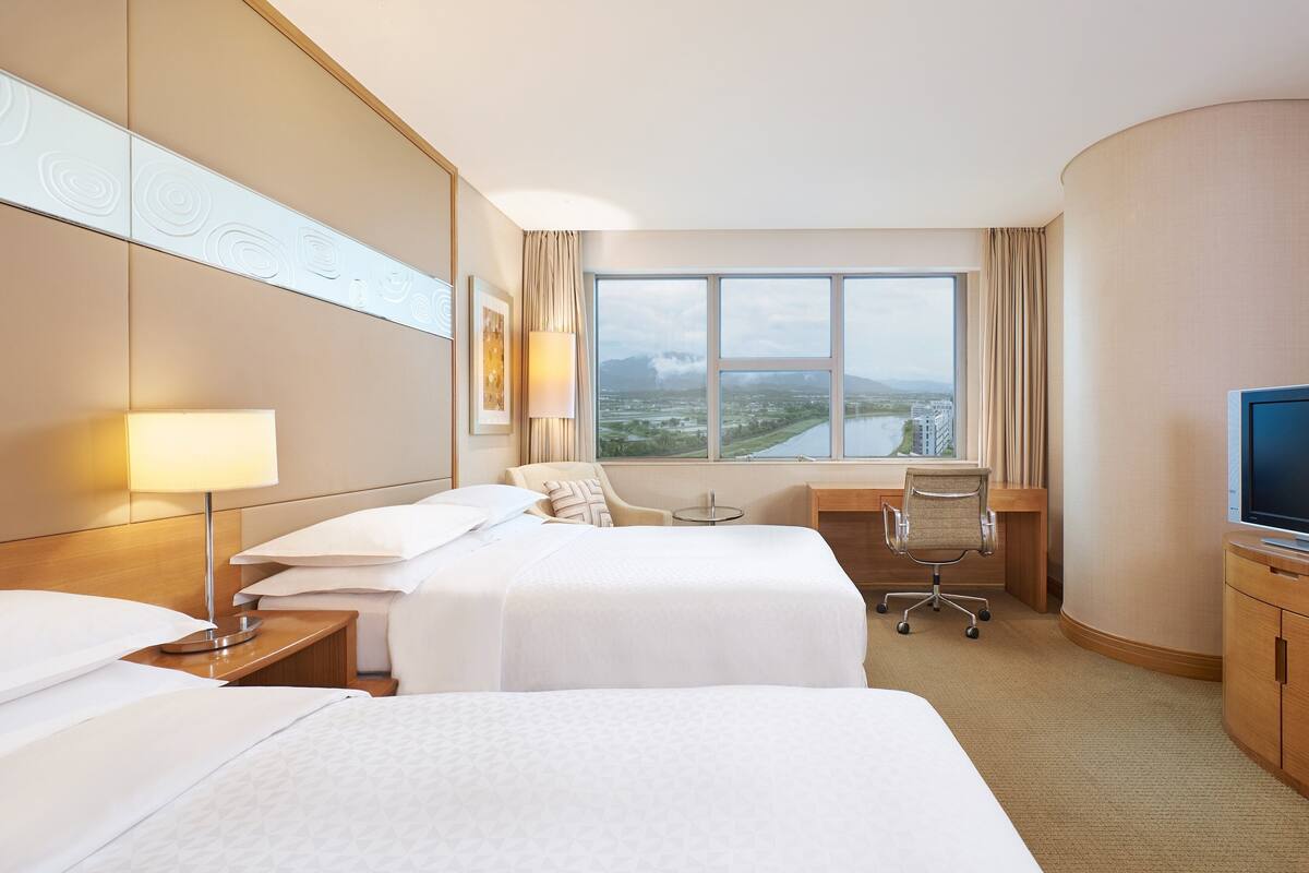 A hotel room with two beds, a desk, a chair, a TV, and a large window with a view of a cityscape.