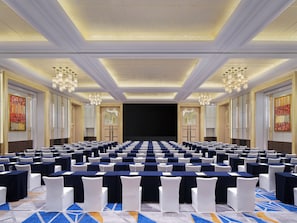 Un salón de conferencias con filas de mesas y sillas, un escenario y candelabros.