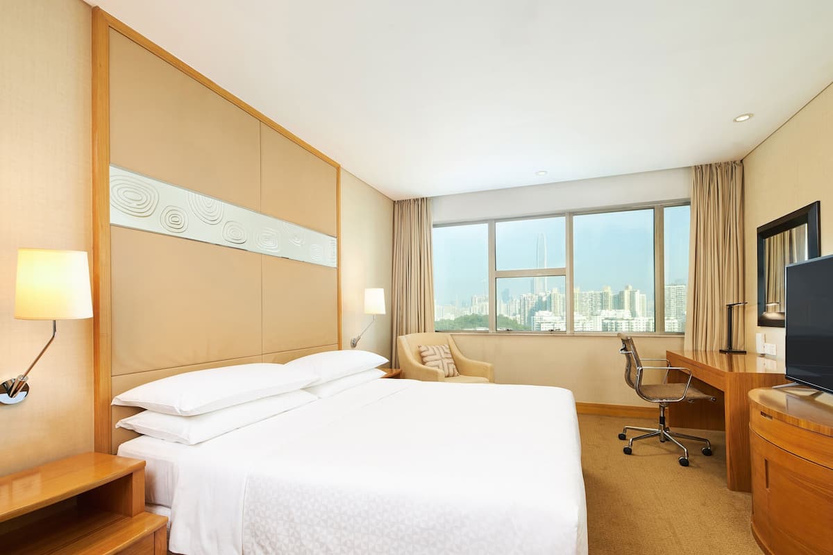 A hotel room with a bed, a desk, a chair, a TV, and a window with a city view.