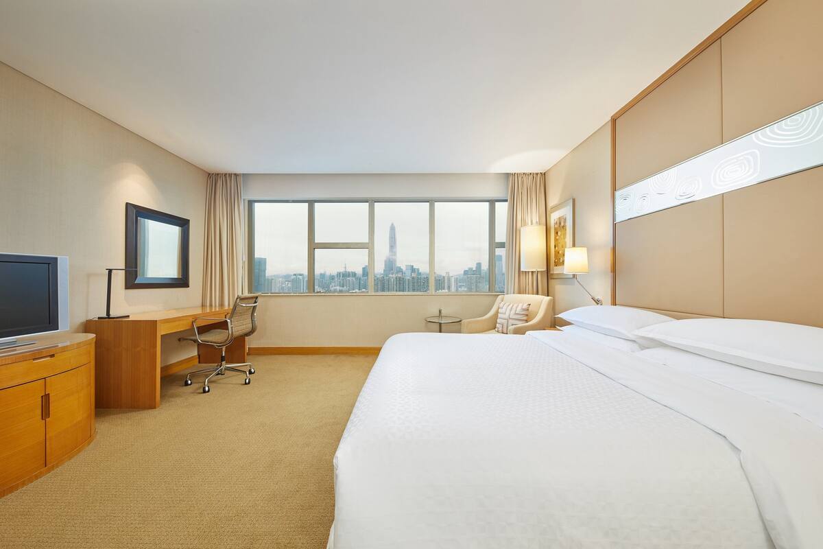 A hotel room with a large bed, a desk, a chair, a TV, and a view of a city skyline through large windows.