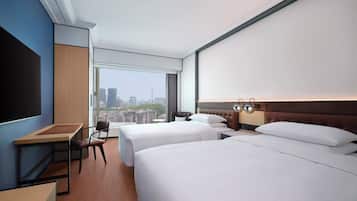 A hotel room with two beds, a desk, a chair, and a large window offering a city view.