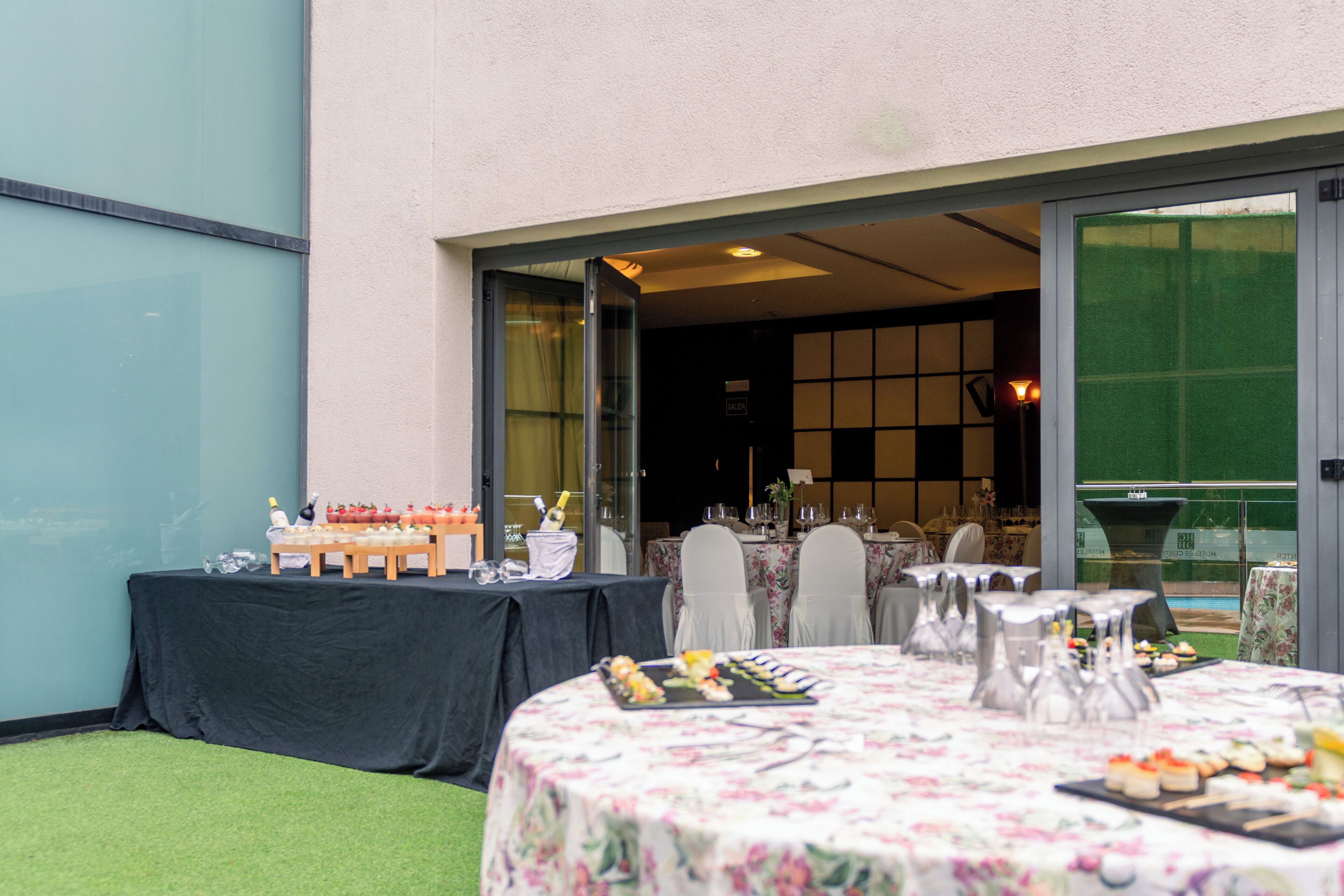 An outdoor dining area with tables set for a meal, a buffet with various food items, and a view of the building's interior.