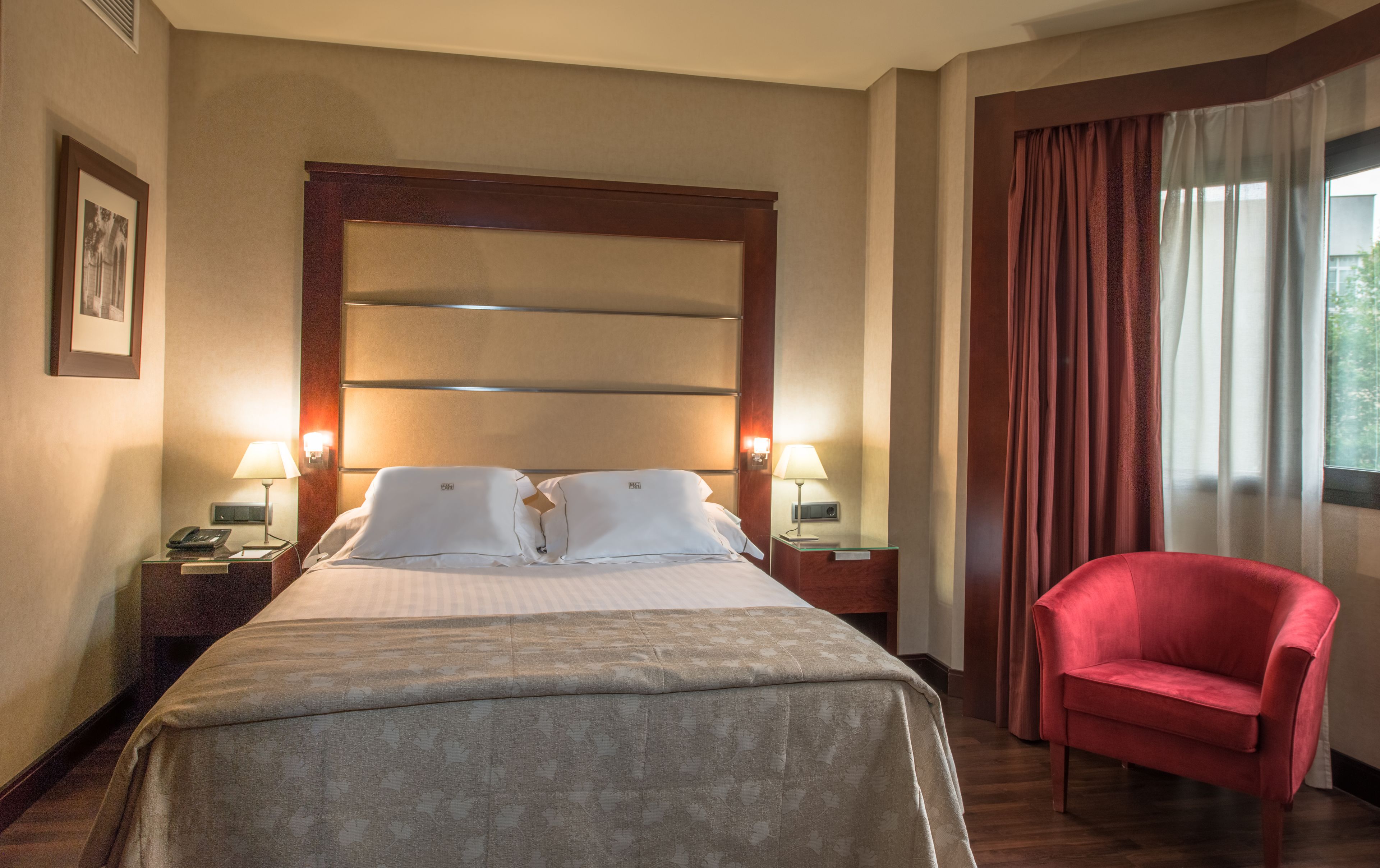 A hotel room with a large bed, two bedside lamps, a red armchair, and a window with curtains.