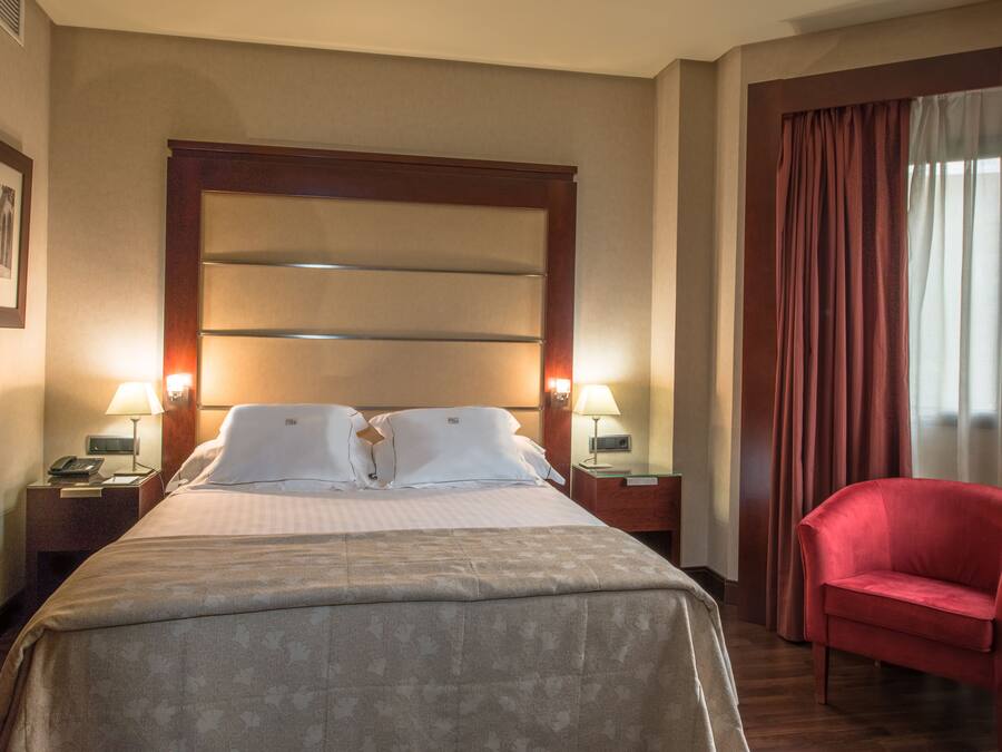 A hotel room with a large bed, two bedside lamps, a red armchair, and a window with curtains.
