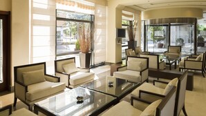 A modern hotel lobby with a seating area featuring sofas and armchairs, a glass coffee table, and large windows offering views of the outdoors.