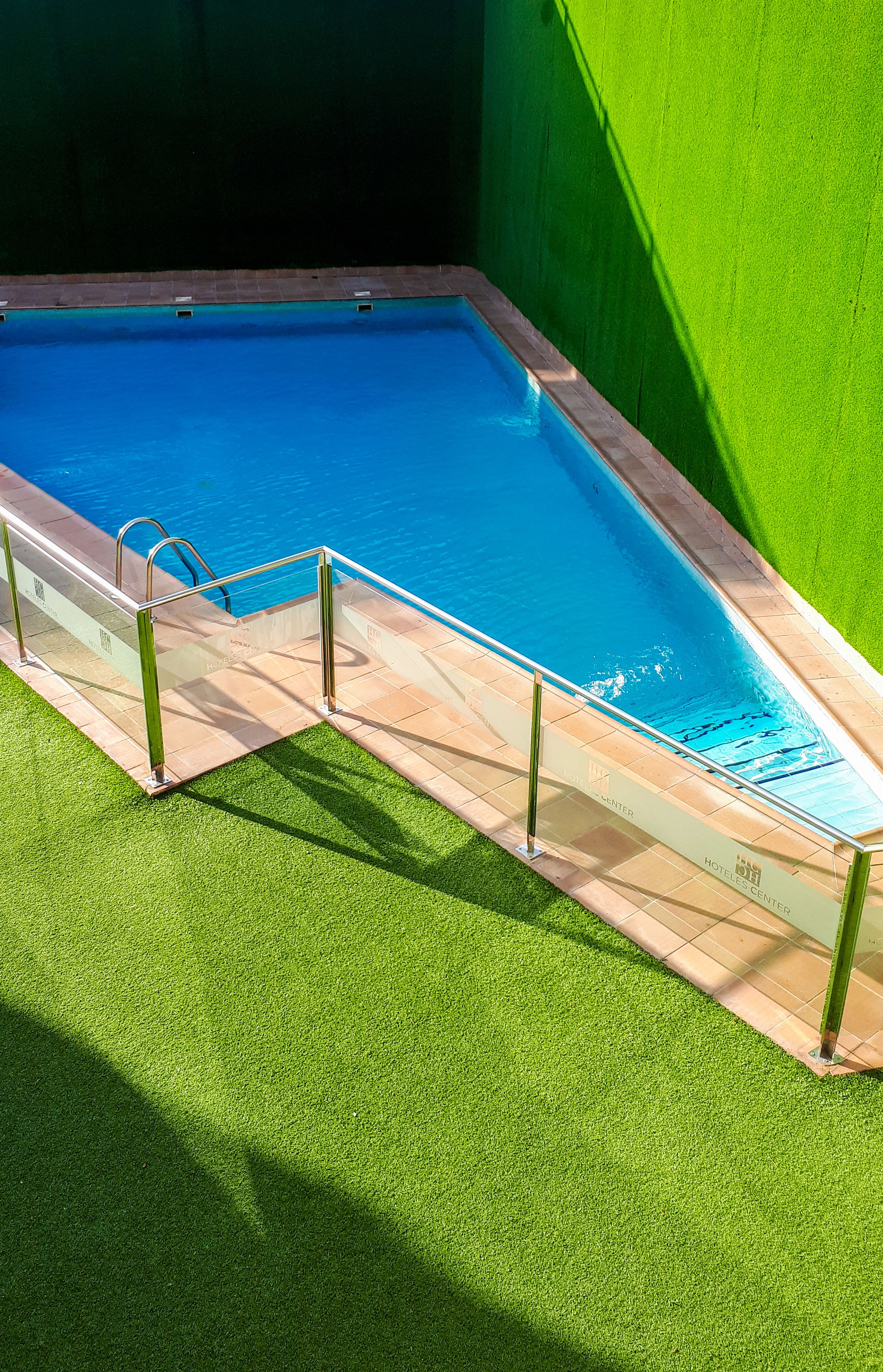 Una piscina con barrera de vidrio y pasamanos metálicos, rodeada de césped artificial y un muro verde.