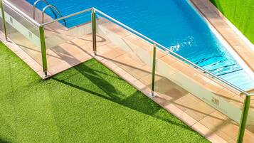 Una piscina con barrera de vidrio y pasamanos metálicos, rodeada de césped artificial y un muro verde.