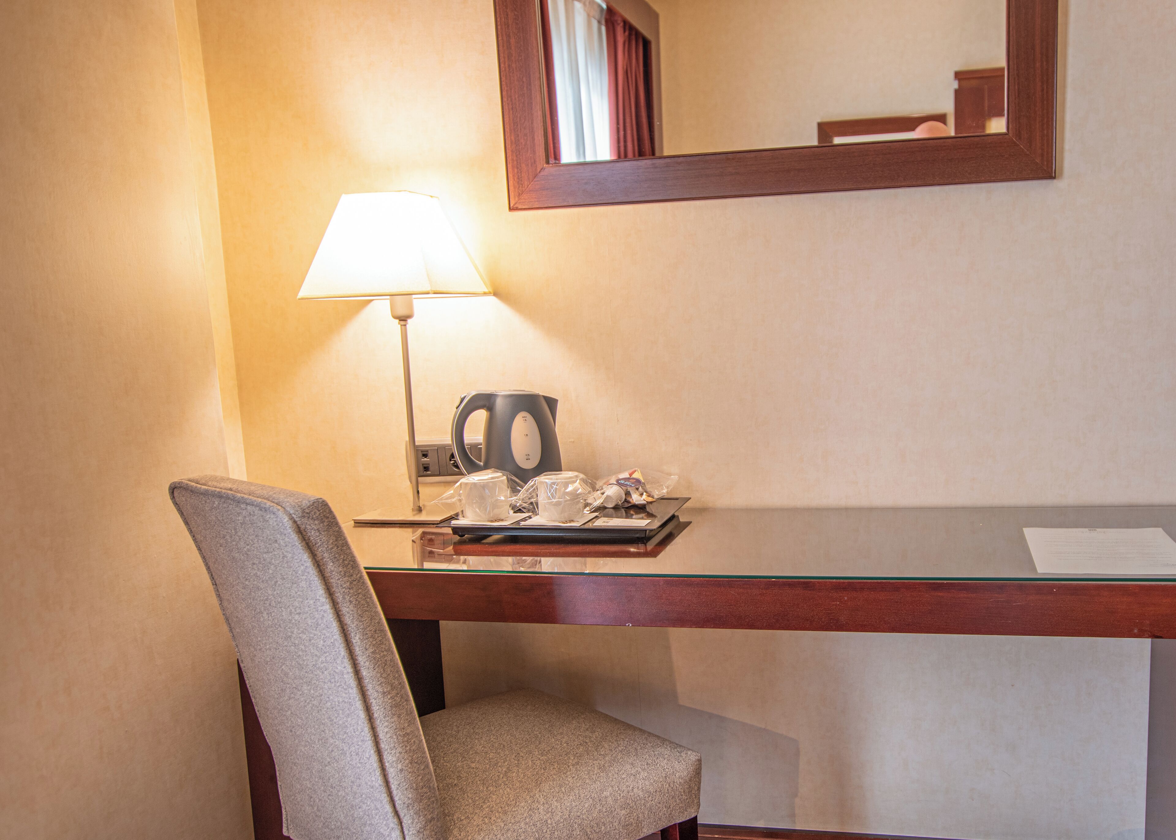 Een hotelkamer met een bureau, een stoel, een lamp, een waterkoker en een spiegel.