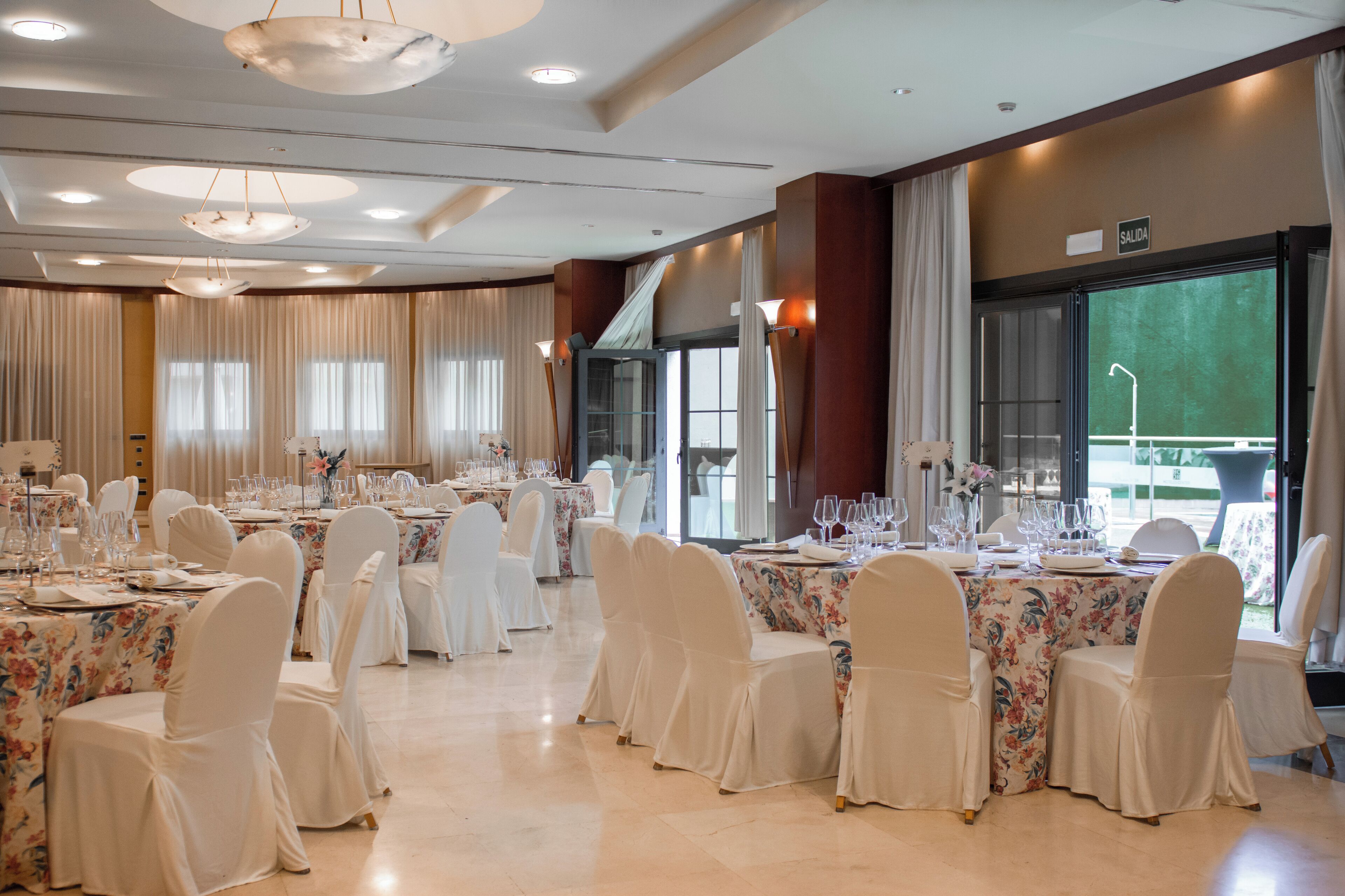 Un salón de banquetes con mesas redondas dispuestas para cenar, sillas blancas y manteles florales.