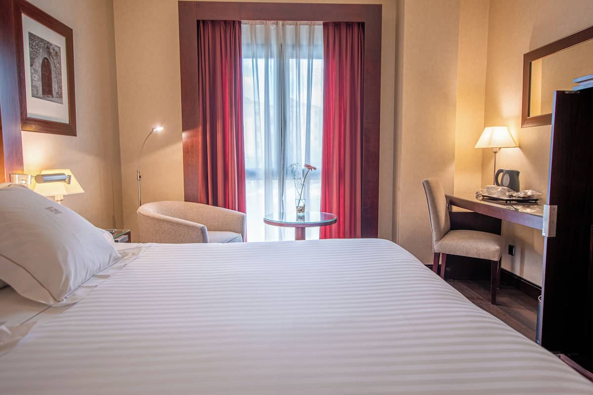 A hotel room with a large bed, a desk, a chair, a small table, and a window with curtains.