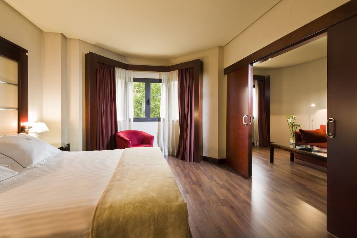 A hotel room with a large bed, a red chair, a desk with a lamp, and a view of greenery outside.