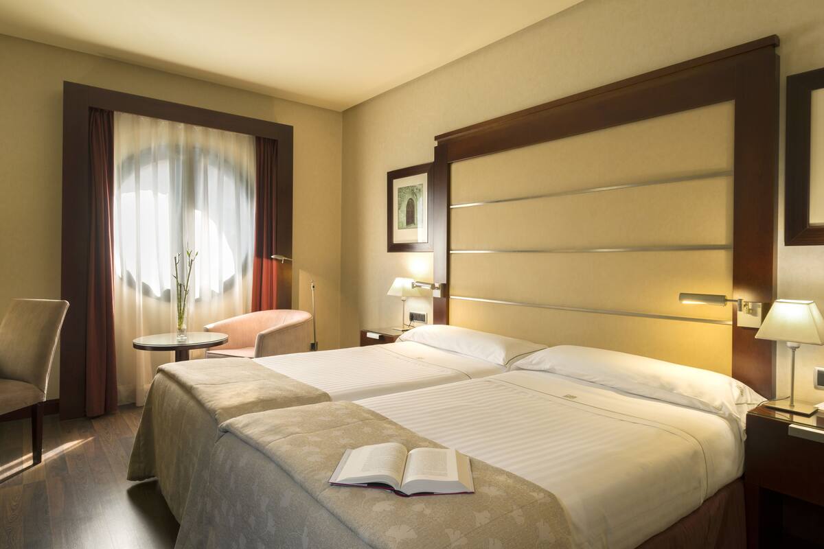 A hotel room with two beds, a chair, a small table, and a lamp.