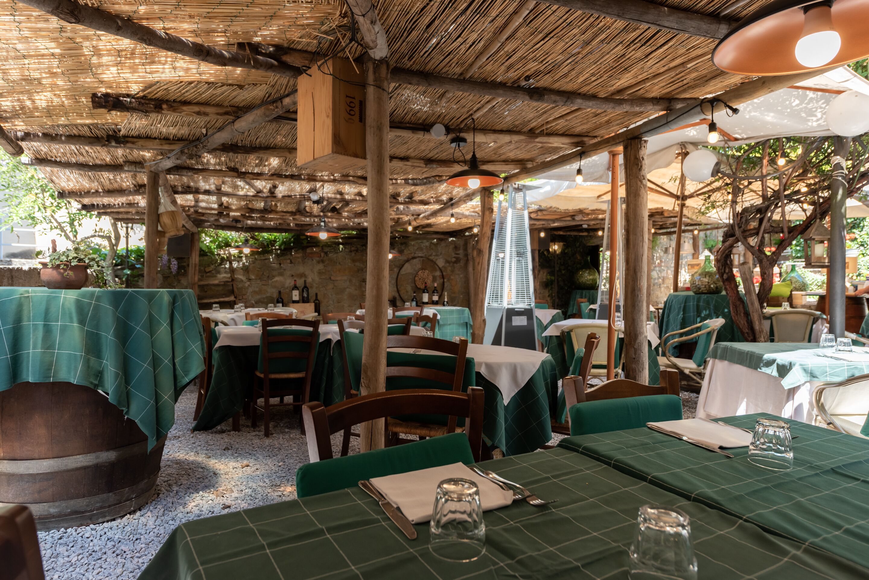 An outdoor restaurant with tables covered in green tablecloths, wooden chairs, and a rustic thatched roof.