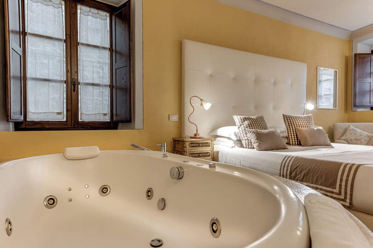 Quarto com uma banheira grande, uma cama com muitas almofadas, mesa de cabeceira com abajur e janela com cortinas.