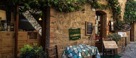 A rustic outdoor seating area with a table covered by a patterned tablecloth, wooden chairs, and a sign indicating the entrance to a garden.