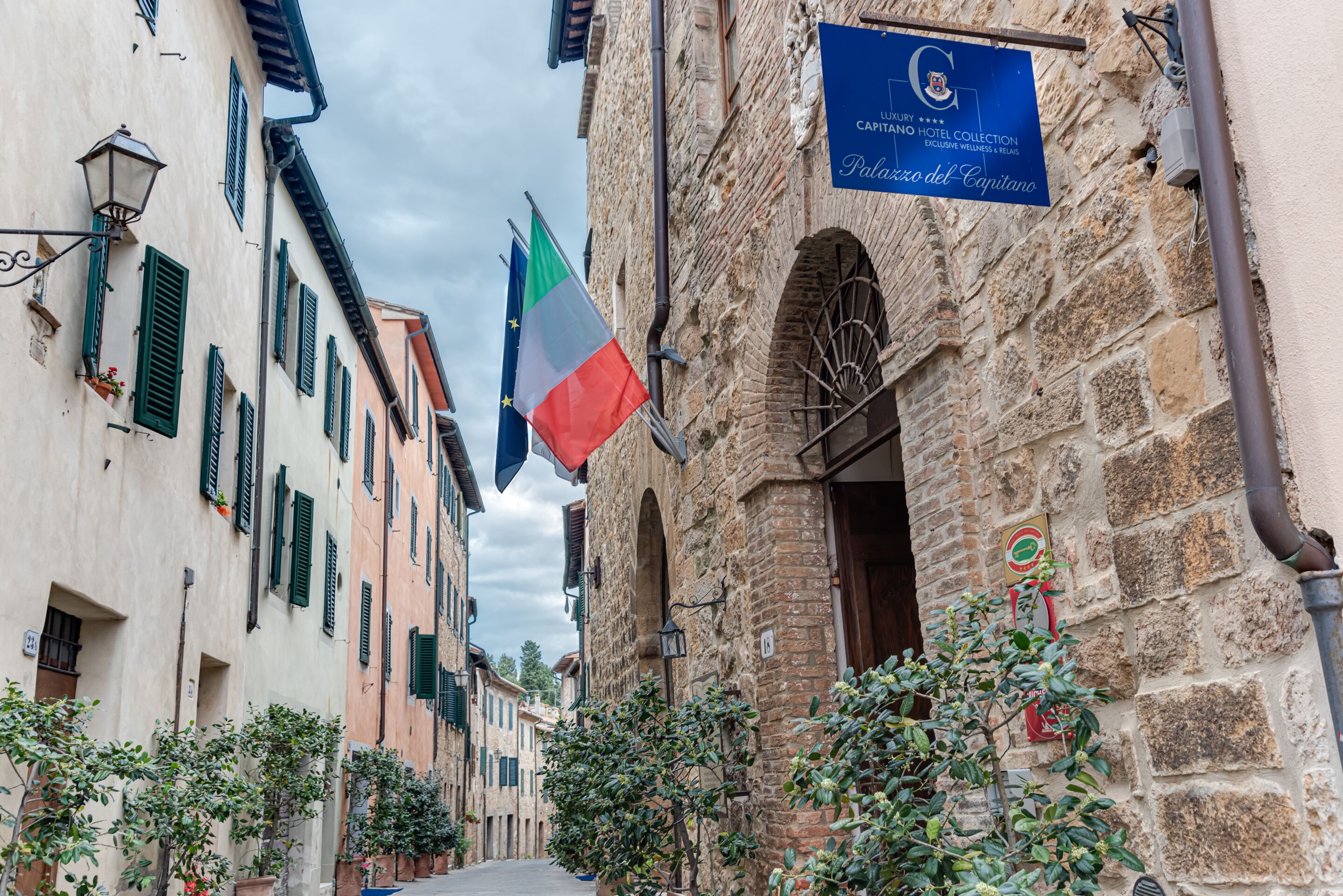 Una calle estrecha con edificios históricos, un cartel que indica Palazzo del Capitano y banderas.