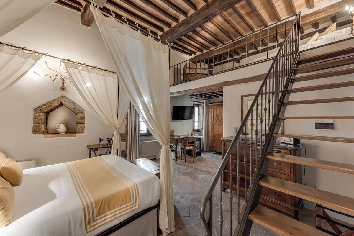 Quarto com cama, uma mesinha, uma cadeira, janela com cortinas, parede de pedra e escada.