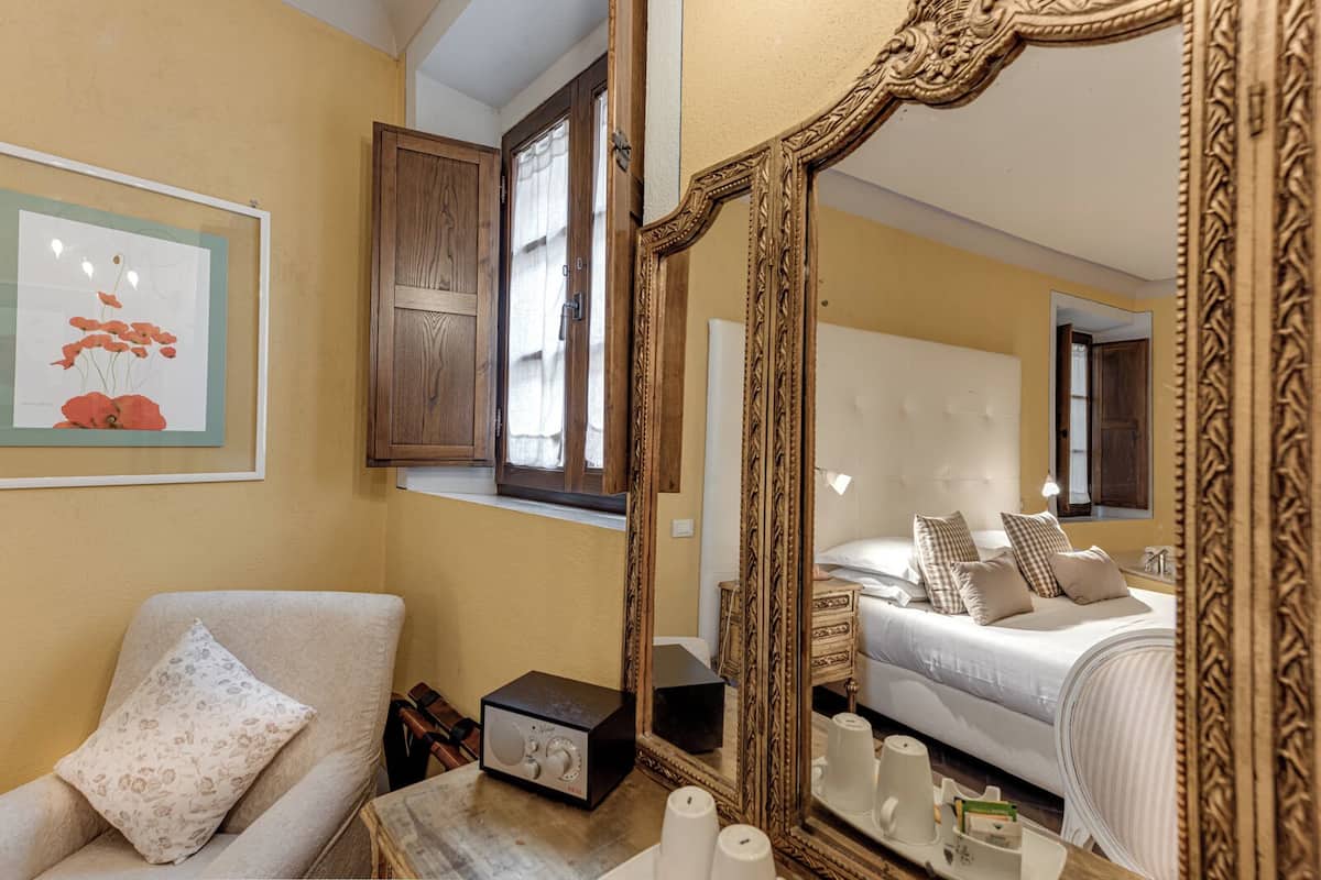 Quarto com um espelho grande, cama com várias almofadas, mesa de cabeceira com um rádio e um quadro na parede.