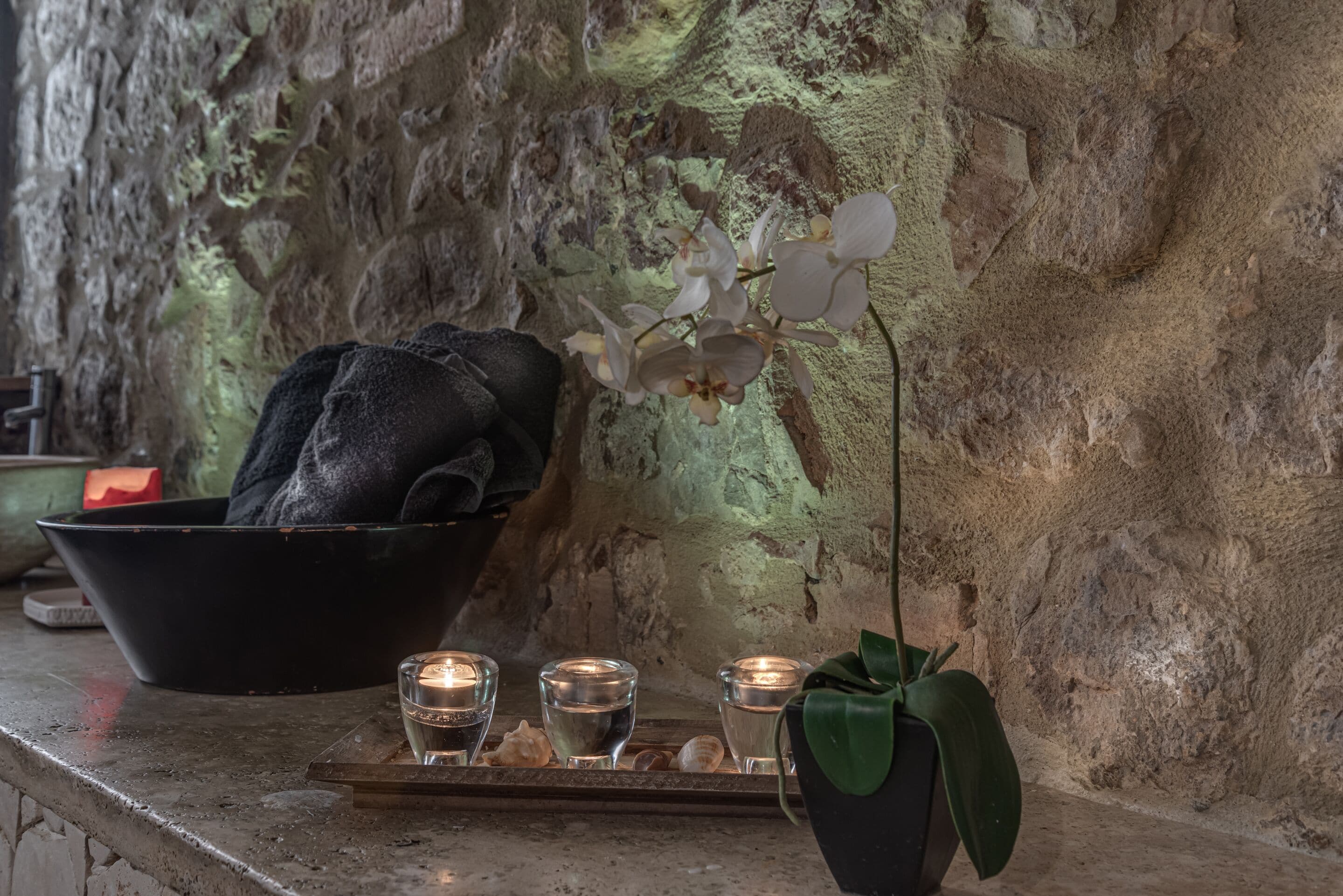 Un mur de pierres sur lequel est posé un bol noir, trois petits verres à liqueur en verre avec des bougies, et une orchidée en pot.