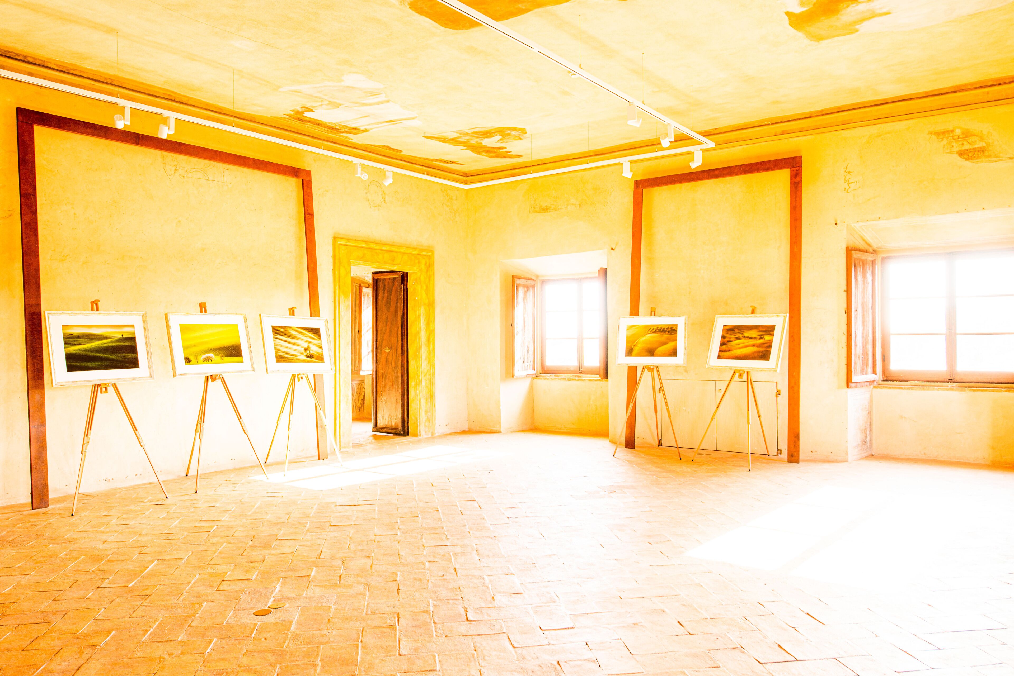 Uma galeria ampla com piso de tijolos e grandes janelas, exibindo várias pinturas em cavaletes.