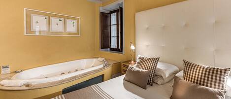 A hotel room with a large bed, a separate bathtub, a bedside table with a lamp, and a window with curtains.