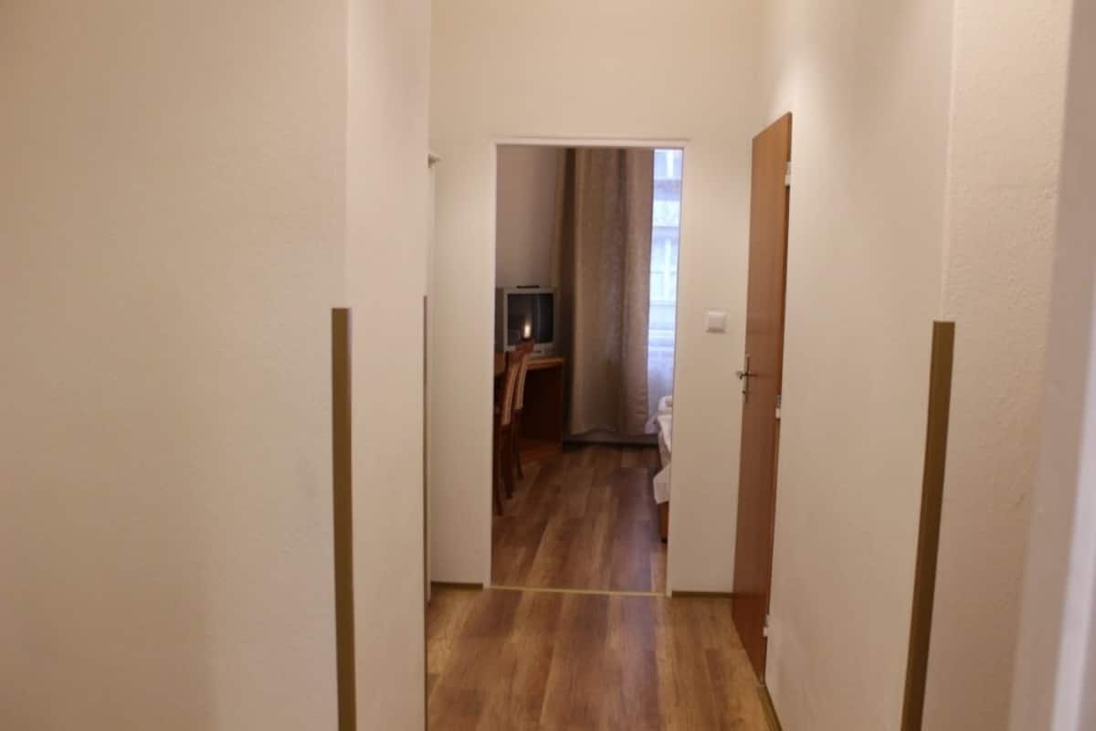 A narrow hallway with wooden flooring leading to a room with a bed and a television.
