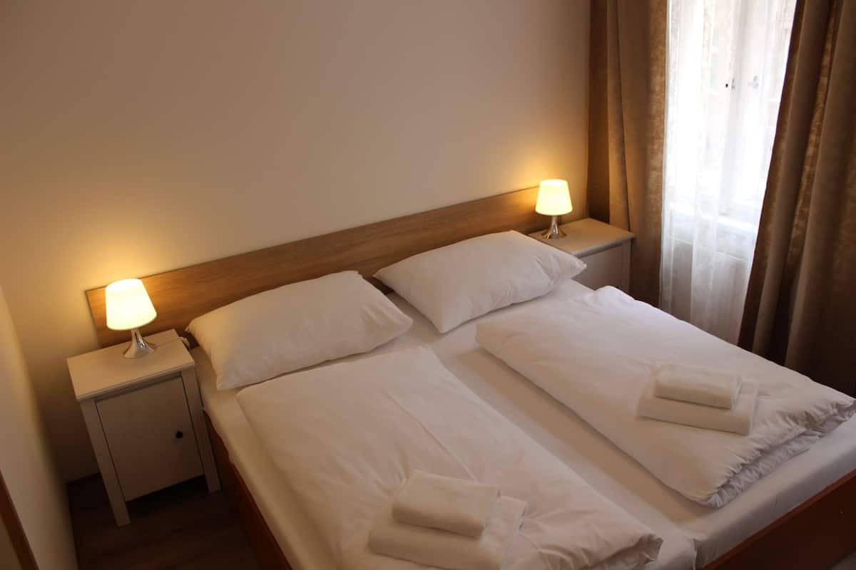 A neatly made double bed with white linens and two bedside lamps, a wooden headboard, and a window with curtains.