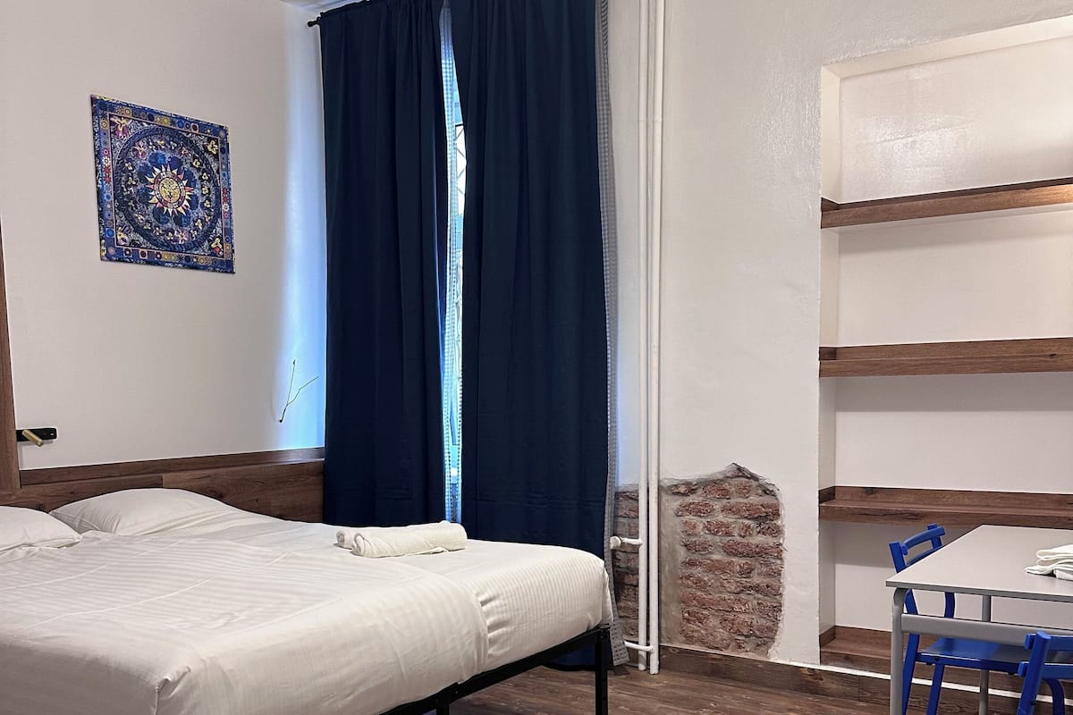 A room with a bed, a table with chairs, a tapestry on the wall, and a brick wall.