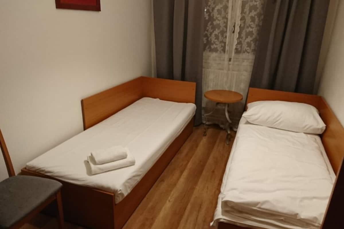A small, single-room hotel bedroom with two single beds, a wooden floor, a small table, and a chair.