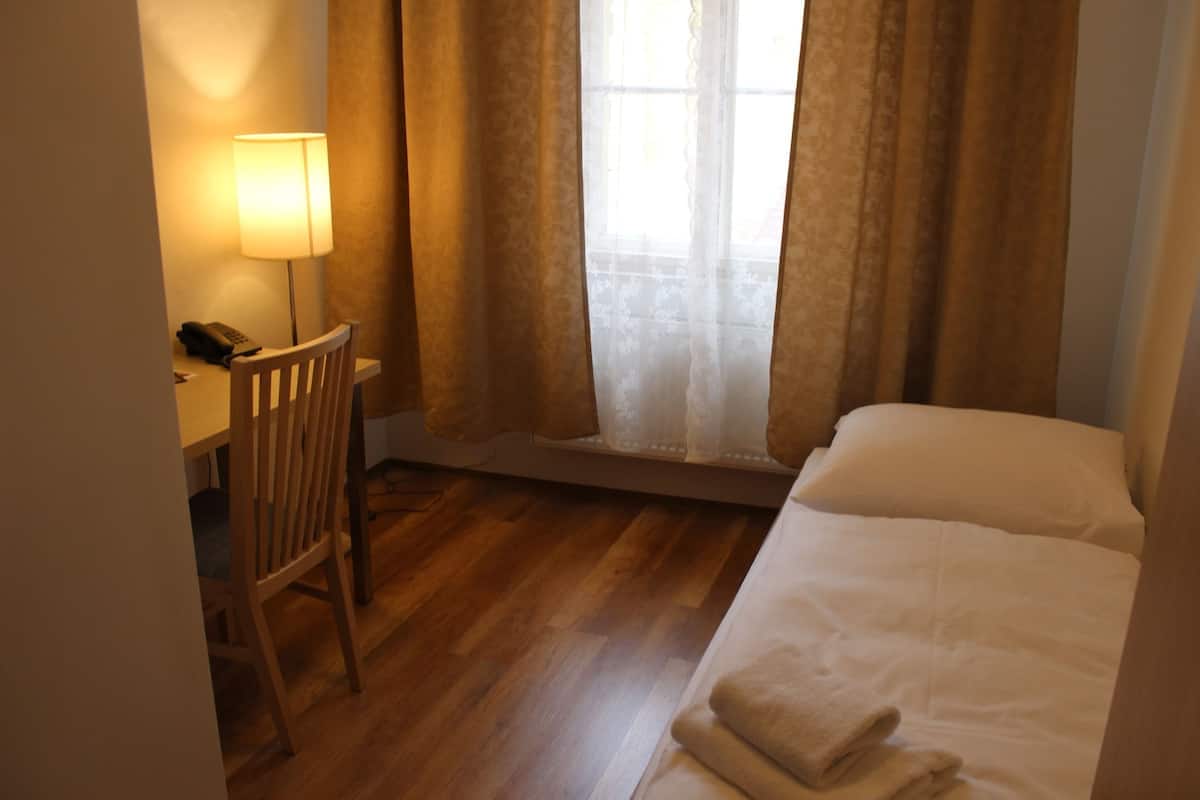 A hotel room with a wooden floor, a desk with a lamp, a chair, a bed with a white comforter, and a window with sheer curtains.