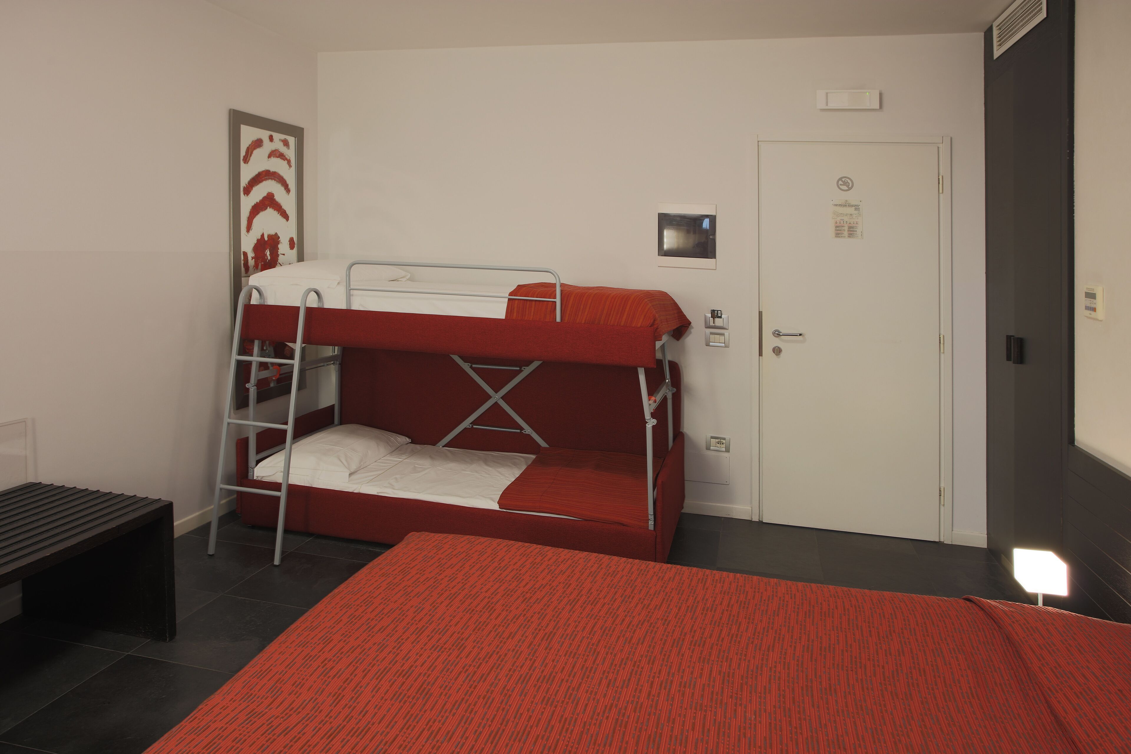 Een slaapkamer met een stapelbed, een rode spreideken, een tafeltje en een deur met een bordje.
