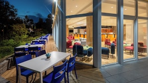 An outdoor dining area with tables and chairs, a wooden deck, and a view into the interior of a building with a bar and seating area. - Hunguest Hotel Helios (Hévíz)