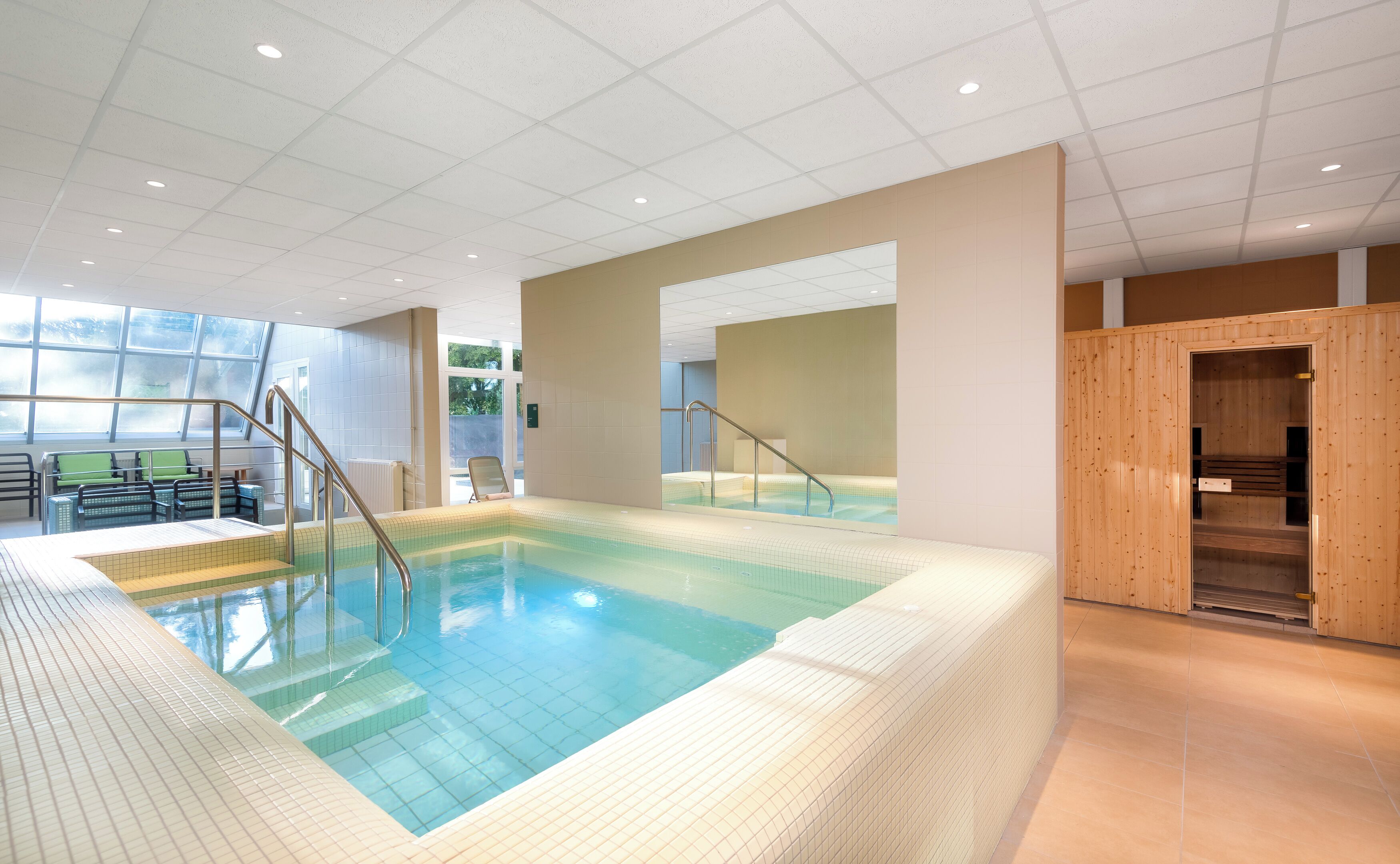 Ein großer, überdachter Schwimmbereich mit gefliesten Stufen und Metallgeländern, umgeben von beigen Wänden und einem Holz-Saunabereich.