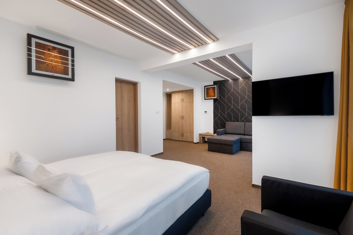 Ein modernes Hotelzimmer mit einem großen Bett, einem flachbildfernseher an der Wand und einer Sitzecke mit Sofa und Sessel.